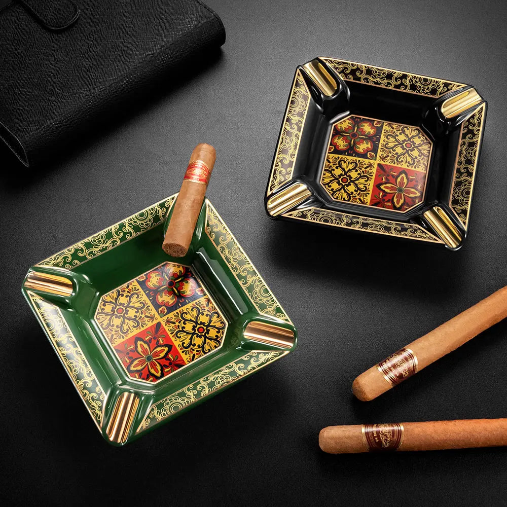 Two decorative ashtrays with intricate designs and cigars on a dark surface.