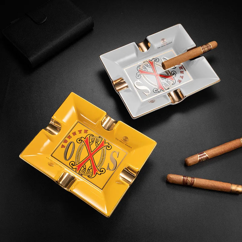 Two ashtrays, one yellow and one white with gold accents, on a dark surface with cigars.